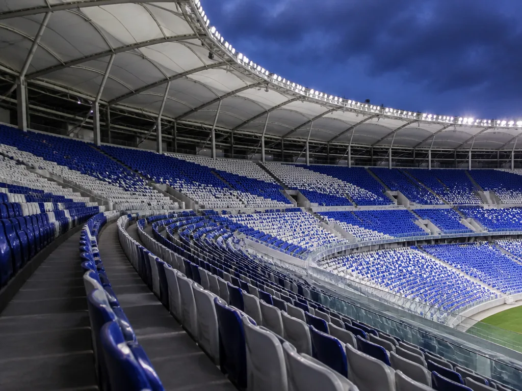 Tomma läktarsektioner i blått och vitt under ett böjt vitt tak på en modern fotbollsarena i kvällsljus
