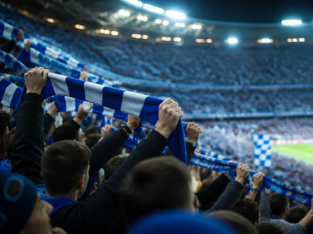 Fullsatt fotbollsstadion med blåvita supportrar som håller upp halsdukar under kvällsmatcher med flodljus