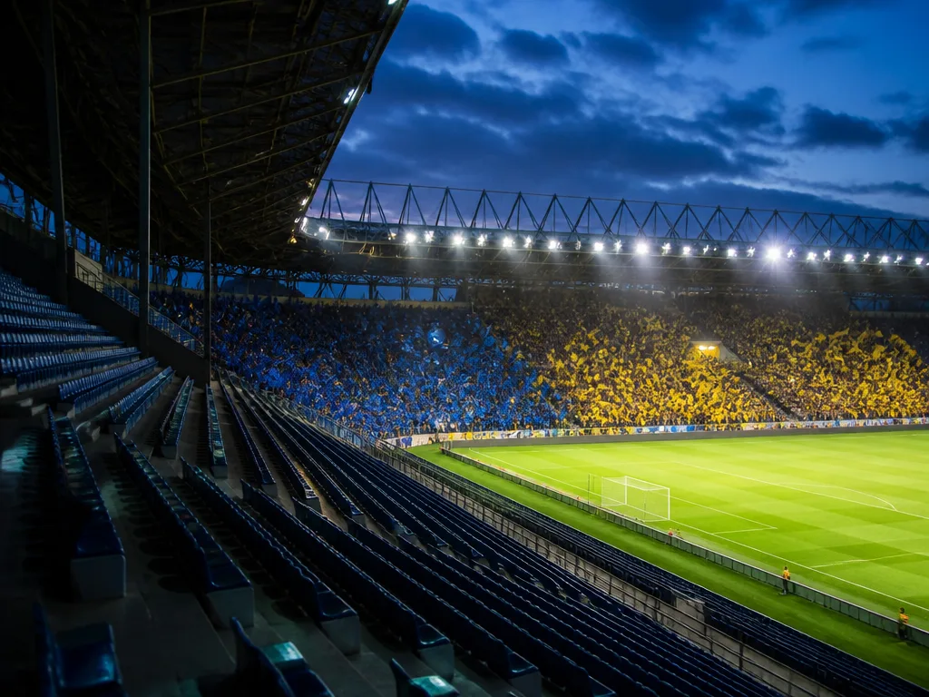 Fotbollsarena i kvällsljus med tänd flodbelysning och tom bortasektion under en Allsvenskan-match