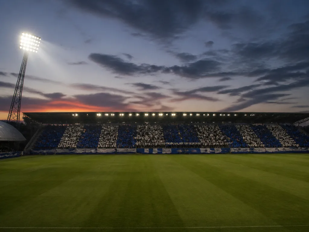 Skandinavisk fotbollsstadion i skymning med blåvita supporterfärger på läktaren och grön plan under strålkastarljus